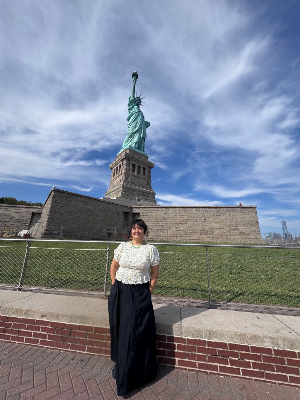 A woman stands on Liberty Island in front of the Statue of Liberty. She is wearing a light patterned short-sleeve blouse and a long dark skirt, standing with her hands in her pockets. The Statue of Liberty rises behind her against a blue sky with thin white clouds.